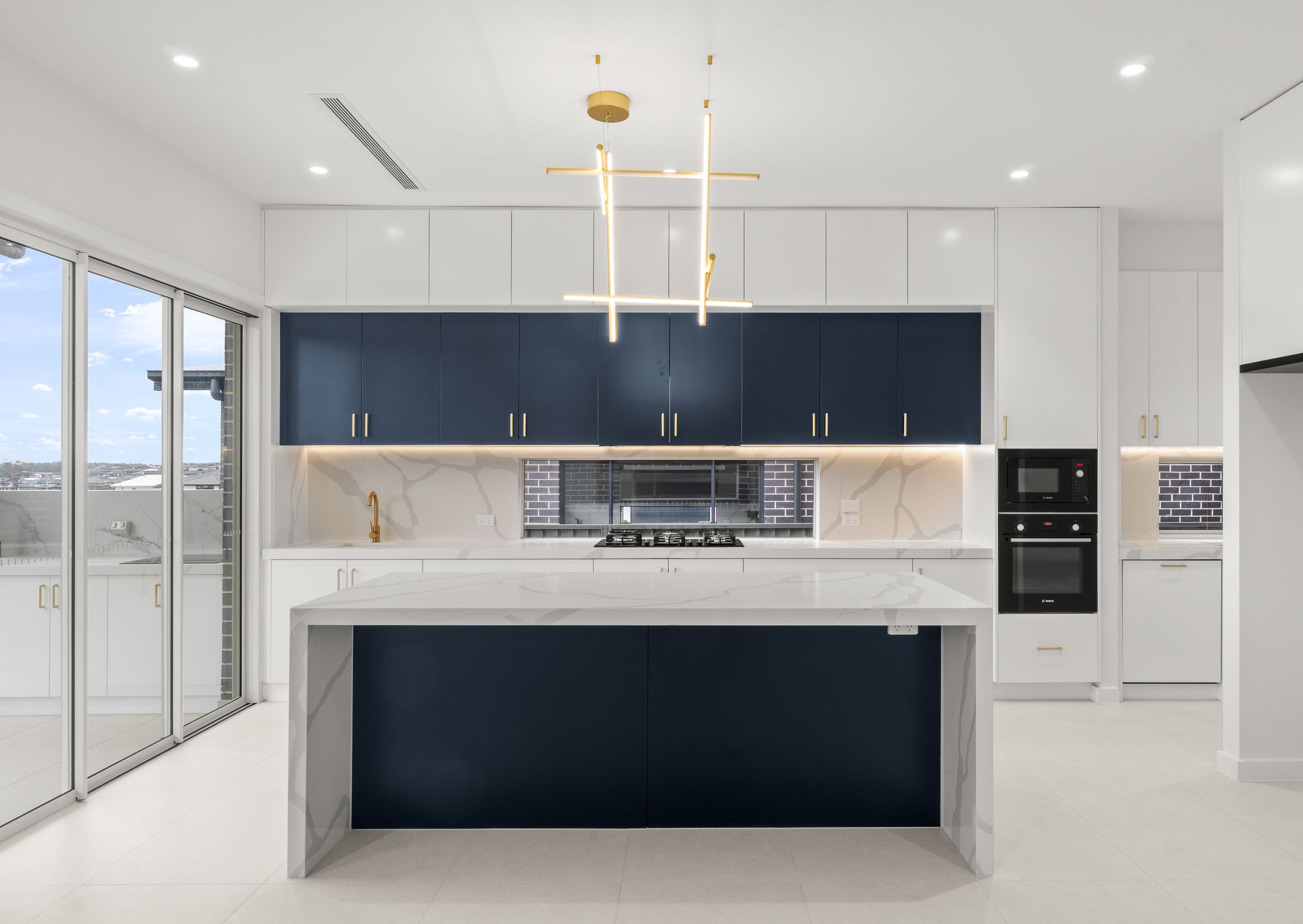 Wide view of a navy and gold kitchen with a marble waterfall island and brass pendant lighting